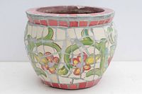Front side showing fragmented tile mosaic of red and yellow flower with green leaves on cream background, with red tile rim. Thick walled clay pottery planter.