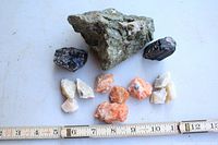 Top view of Canadian rock and mineral specimens with ruler for scale showing greenish rock, dark crystals, orange and white minerals.