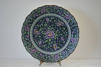 Front view of a large round porcelain charger plate with scalloped edges and intricate floral pattern in purple, green, and white on a dark blue background.