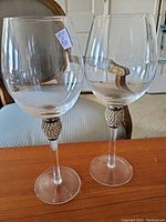 Two brandy snifter glasses with clear bowls and decorative beaded elements on the stems, shown on a wooden table with a chair in the background.