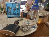 Wide shot showing all barware items on white porcelain tray with silver trim, Evian Millennium water bottle, wine glass charm set, round stone coasters, hip flask, cocktail strainer, and various bar accessories.