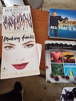 Photo showing several books including 'Fountain Pens', 'Making Faces' by Kevyn Aucoin, 'All That's Golf', and 'Couleurs locales' arranged on a wooden table and chair.