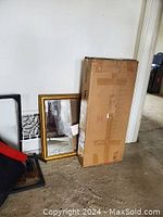 Photo showing a large cardboard box protecting the C shape end table, placed upright on the floor next to a gold framed mirror leaning against the wall.