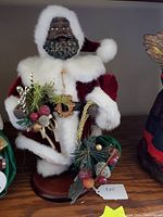 12-inch black Santa figurine wearing red velvet and white trim holding a decorative arrangement with berries and greenery.