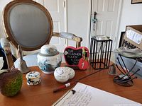 Tabletop displaying all decorative items including ceramic heart, various trinket boxes, decorative apple, small brass bell, candle snuffer, and oil burner arranged on wooden surface with a chair in the background.