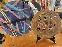 Two ceramic decorative plates standing on display stands on a table. The left plate is large with an abstract blue, black, yellow, purple, and light blue design. The right plate is tan with an ethnic painting of three women carrying vessels.