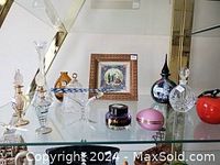 Display shelf with various glass collectible items: crystal and glass perfume bottles, small glass decorative objects including an apple and Christmas bell, a small round pink container, and framed Asian-themed picture.