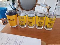 Four Hollywood Park Gold Cup water glasses with yellow and black graphics, placed next to a McCafe cup and a paper with writing.