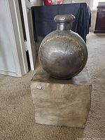 Front view of round metal jar on metal cube stand on carpeted floor with partial room background.