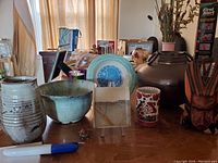 Photo showing multiple decorative artifacts: ceramic vases and bowls, plates, a wooden sculpture, brass vessel, leather pouch, and a decorative ceramic cup.