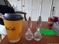 Two clear crystal perfume bottles with pointed stoppers on a wooden table with a McCafe coffee cup for scale.