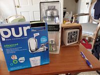 Photo showing various kitchen items on a table: Pur water filtration system in box, Rabbit cocktail mixer, vintage clock, and a blue pen.