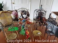 Photo showing 10 miniature decorative metal chairs in various colors and designs, some holding dolls and cushions, set against a white wall with miscellaneous background items.