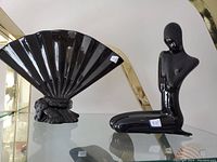 Photo showing a black ceramic folding fan sculpture next to a black ceramic abstract human statue, both on a glass shelf under display lighting.