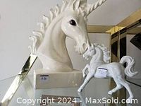 Photo of large white ceramic unicorn head sculpture and small white ceramic unicorn figurine together on a glass shelf.