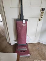 Photo shows Hoover Elite 350 maroon upright vacuum cleaner leaning against a door. Extra vacuum bags are in the background on the floor.