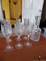Set of 6 crystal stemmed glasses (3 champagne flutes and 3 wine glasses) and a crystal wine decanter displayed on table near an open door.