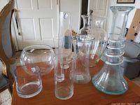 Photo showing eight clear glass vases on a table, various shapes including round, cylindrical, and wide base with rings.