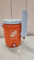 Full view of orange Home Depot water cooler showing front, lid, spigot and attached cup dispenser with cups inside.