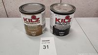 Two new sealed cans of Kilz primer, sealer, stain, and odor blocking products on a white surface.