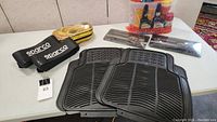 Overview of car care accessories lot on table including Sparco seatbelt pads and cushions, black rubber floor mats, yellow coiled hose, and car care bottles