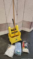 Yellow commercial mop bucket with wringer and wheels together with three mop heads in original packaging placed in front.
