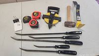 Photo showing four hand scrapers with black handles, a hammer with a wooden handle, a pry bar with yellow and black handle, and three measuring tapes.