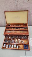 Open wooden storage case showing three rows of assorted rotary tool bits and a drawer below with compartments holding accessories and polishing compounds.