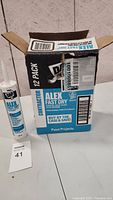 Opened box with Dap Alex Fast Dry caulk tubes visible, showing 12 pack labeling.
