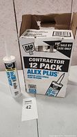 Opened contractor box with 12 tubes of DAP Alex Plus acrylic latex caulk plus silicone and one tube outside the box showing white caulk tube with nozzle.