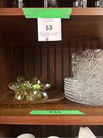 Shelf with stacks of clear glass dessert plates and crystal bowls, two yellow crystal flower-shaped napkin rings on left, under labeled shelf #53