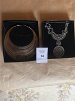 Two metal necklaces in boxes, one gold-tone multi-hoop collar style and one silver-tone ornate pendant necklace.