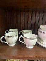 Four white china cups with silver pattern on rim and curved handles on a wooden shelf.