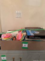 Box with assorted office supplies including sticky notes, paper, and black Pendaflex hanging folders.