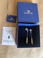 Pair of silver-tone Swarovski earrings displayed in original blue jewelry box, showing the front and back of earrings with branded tag nearby.