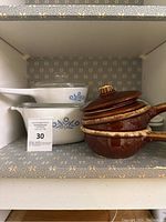 Shelved view showing two CorningWare Blue Cornflower casserole dishes with glass lids beside three brown ceramic handled casseroles with lids.