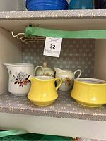 Four vintage ceramic creamers and sugar bowls on a shelf with floral wallpaper background. Two white with floral patterns, two yellow pieces.