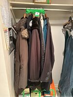 Seven women's coats hanging on hangers on a metal rack, varying in color and material. Mostly long coats, with functional details such as a fur-trimmed hood on one.