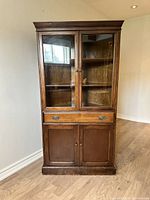 Front view of the antique corner cabinet showing the glass doors, drawer with two handles, and bottom cupboard with wooden doors.
