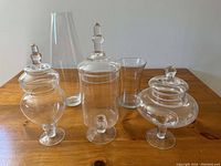 Front view of three apothecary jars with lids and two glass vases on a wooden surface under good lighting.