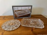 Photo showing two crystal trays on a wooden table, one rectangular in a box (new) and one round tray.