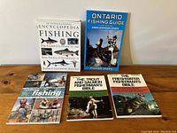 Five fishing books displayed upright and lying down on a wooden surface showing front covers.