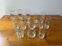 Set of 13 branded beer and whisky glasses arranged on wooden surface, including Stella Artois, Becks, Corona, Hoegaarden, and Canadian Club glasses.