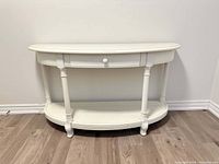 Front view of white half moon console table with drawer and bottom shelf on wood flooring against wall.