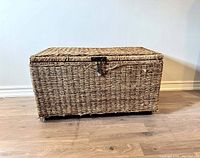 Closed view of large wicker chest, showing rectangular shape, woven wicker surface, and latch on front.