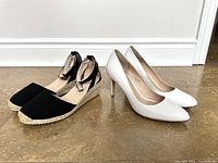Front and side view of black wedge sandals and white high heel shoes displayed on floor against white wall.