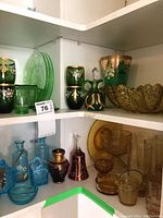 Photo of multiple colored Depression glass pieces including emerald green hand-painted glasses, blue glassware, and amber depression glass items organized on white shelving.