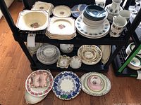 Multiple assorted plates, bowls, and dishes displayed on two black shelves. Includes floral and pattern plates, various colors and designs.