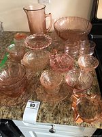 Photo showing a group of various peach/soft pink colored glass pieces including bowls, glasses, plates, a lidded dish, and a pitcher arranged on countertop.