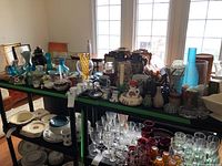 Wide view of two tables showing assorted decorative ceramics, glassware, and vases placed near windows with natural light.